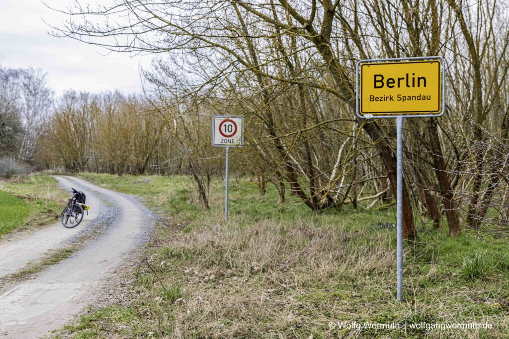An einem Feldweg, Waldweg steht ein Ortseingangsschild Berlin-Spandau. Stückchen weiter Geschwindigkeit auf 10 kmh begrenzt.
